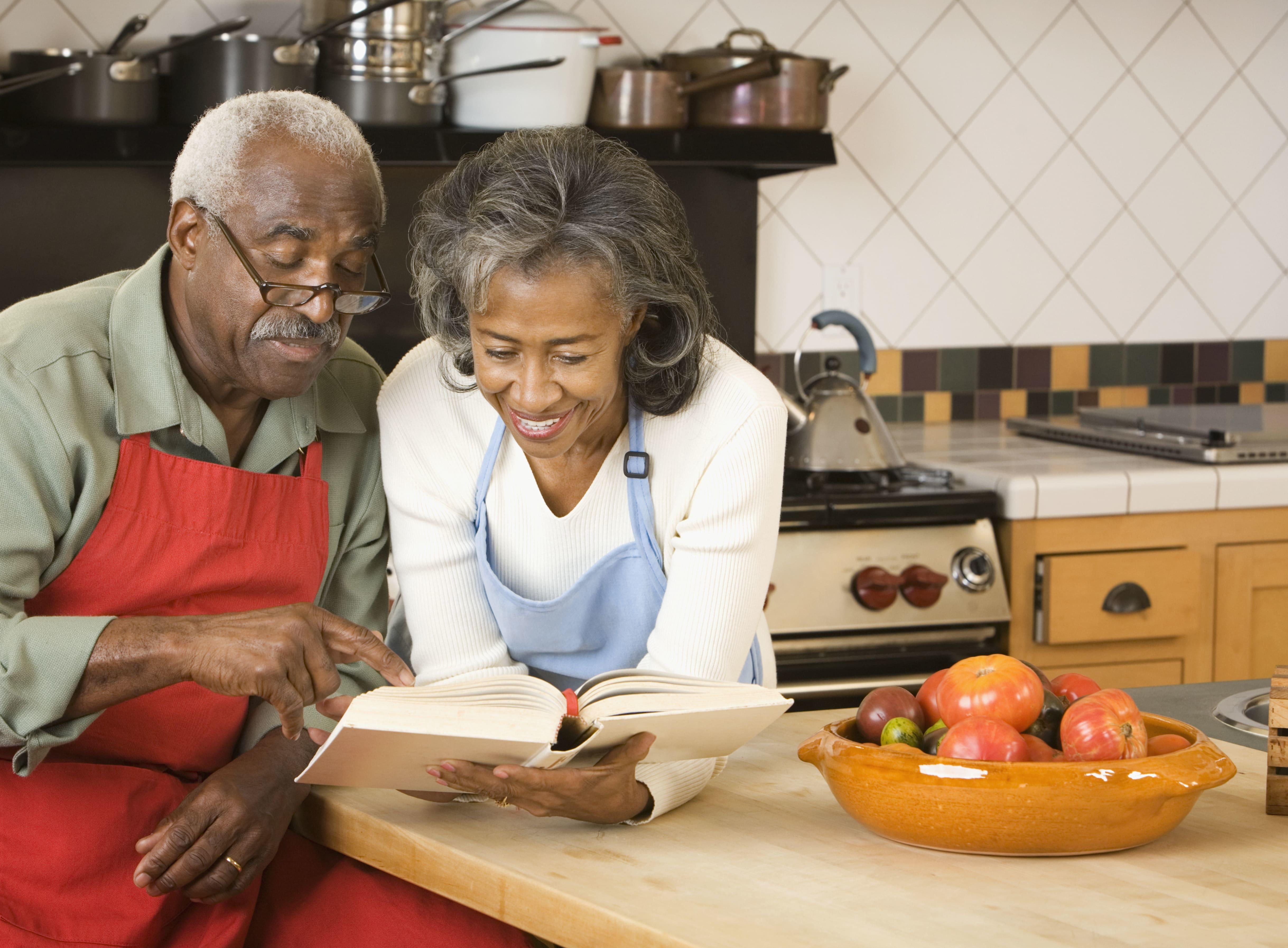 Man wearing glasses sitting next to a woman and looking at a cookbook together at a kitchen counter with fresh vegtables in a bowl