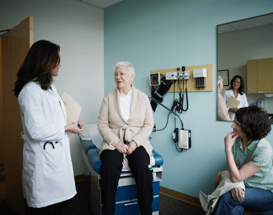 Female patient with glasses sitting on exam bench in doctor’s office talking to female doctor while her female caregiver sits next to her