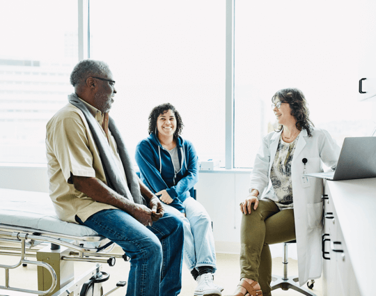 Smiling female doctor sitting on stool talking to male patient and smiling female caregiver in doctor’s office